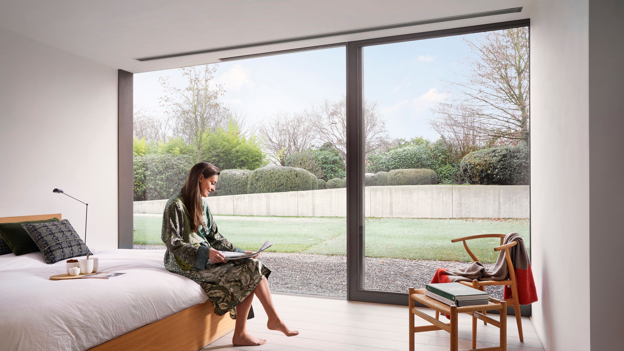 Une femme assise sur le bord de son lit lisant le journal, sa boisson du matin prête et la gainable assurant une température confortable dans la chambre à coucher.