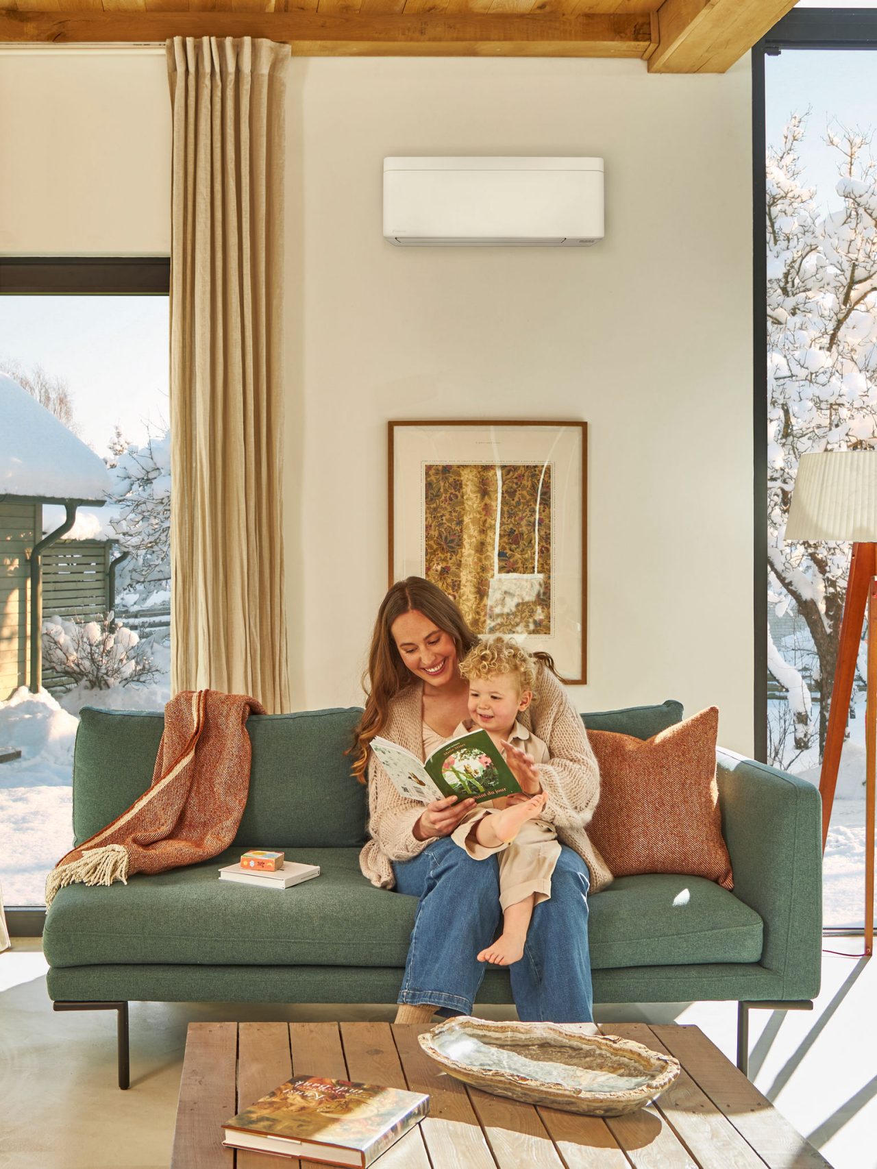 Mère assise sur le canapé sous une pompe à chaleur air-air murale, lisant un livre à son enfant assis sur ses genoux